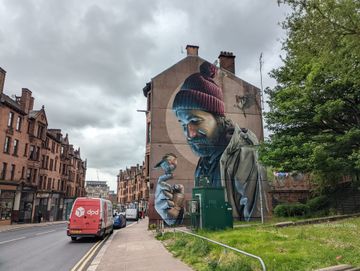 De um lado, vemos uma fileira de prédios avermelhados do primeiro plano até o fundo. Ao centro, outra fileira de prédios, com a lateral coberta por um grafite de um homem que tem um passarinho pousado sobre sua mão. À direita, parte de uma árvore. Dia nublado.