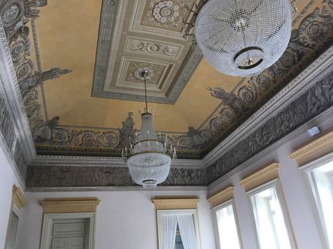 Parte da área interna do teto de um salão. Paredes brancas, teto amarelo e toda a borda em pinturas monocromáticas em tons de cinza, imitando relevo. Dois lustres pendem do teto.