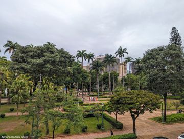 Em primeiro plano, vê-se uma praça cheia de árvores, palmeiras e alguns caminhos no meio, cercados de vegetação cortada em paralelepípedo. Ao fundo, um edifício cinza coberto de linhas horizontais na fachada.
