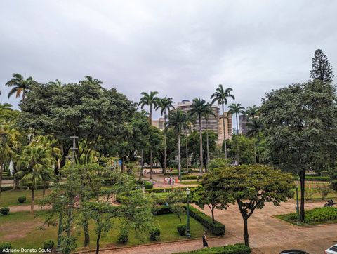 Em primeiro plano, vê-se uma praça cheia de árvores, palmeiras e alguns caminhos no meio, cercados de vegetação cortada em paralelepípedo. Ao fundo, um edifício cinza coberto de linhas horizontais na fachada.