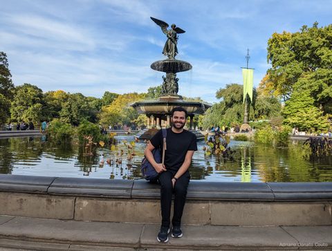 Um rapaz de preto posa em frente a uma fonte circular, com muita vegetação no espelho d'água. Ao centro na fonte, um monumento com um anjo no topo.