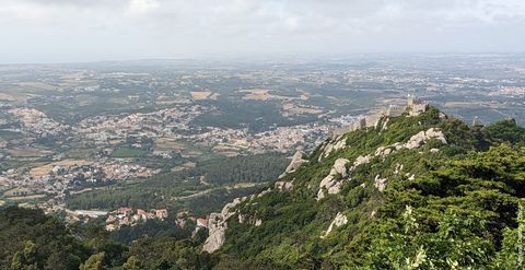 'A partir de uma montanha mais alta, vemos o topo de outra montanha à direita, com muralhas de pedra. Ao fundo, diversos agrupamentos de construções no horizonte.'