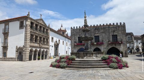 'Sob um céu azul com nuvens, vemos uma praça de piso de pedra. Ao meio, um chafariz (sem água). A praça é ladeada por algumas construções bem antigas.'