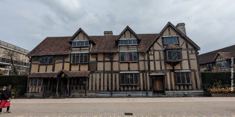 Vemos aqui a fachada de uma casa com paredes em tons de bege e estrutura de madeira escura. Alguém caminha a partir do ponto esquerdo da imagem.