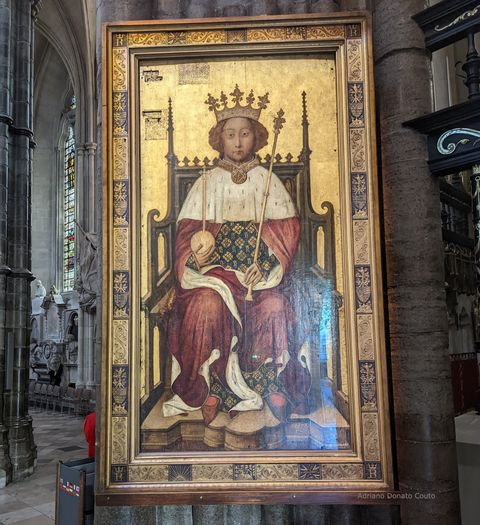 Dentro de uma igreja gótica, vemos um quadro de moldura e fundo dourados, que apresenta o retrato de um rei em seu trono de coroação. O monarca segura o cetro e a orbe. Seu manto é vermelho e a roupa por baixo parece ser em azul escuro com detalhes em dourado.