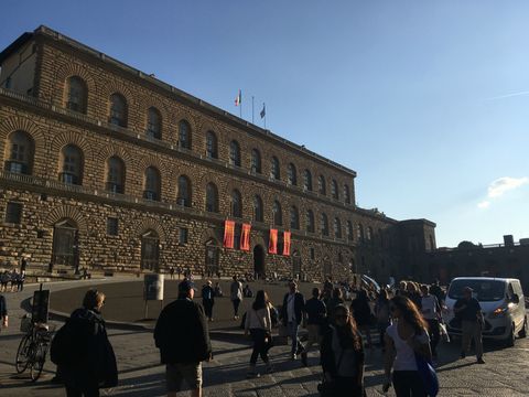 Um enorme palácio com tons entre o cinza e o marrom, construído em formato bem reto. à sua frente, um grupo de pessoas caminhando em diferentes direções.