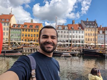 Rapaz moreno de cabelo preto e camiseta azul escuro tira selfie em frente a um canal, com uma sequência de barcos e prédios coloridos ao fundo.