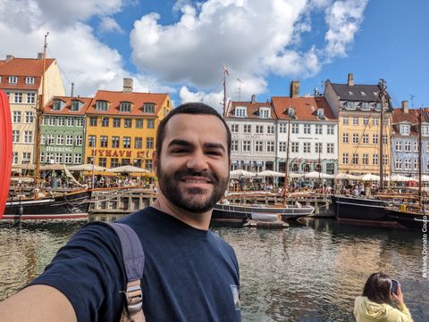 Rapaz moreno de cabelo preto e camiseta azul escuro tira selfie em frente a um canal, com uma sequência de barcos e prédios coloridos ao fundo.