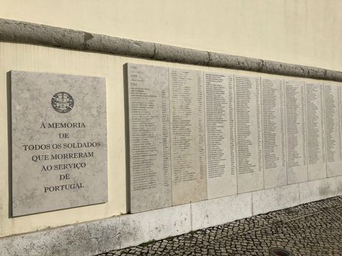 Visão em 45 graus (na horizontal) de uma parede. Esta parede carrega uma placa com os dizeres "À memória de todos os soldados que morreram ao serviço de Portugal".  Do lado direito, várias placas com nomes dos referidos soldados homenageados.