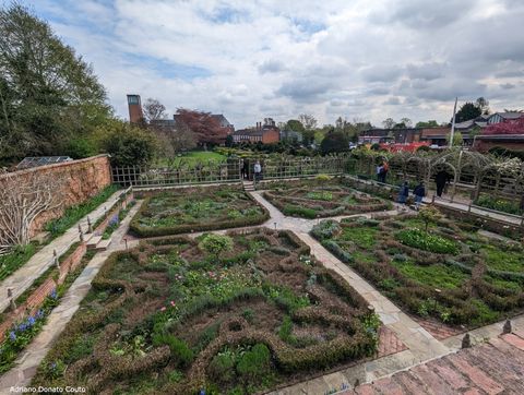 Temos um jardim geométrico, com plantas podadas que formam desenhos em quatro seções quadradas. A cor predominante é verde. O céu está nublado.