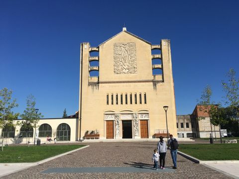 Um casal e uma criança pequena caminham por uma via de pedra em direção a uma igreja cor creme, que possui um detalhe em relevo no topo. Céu azul sem nuvens.