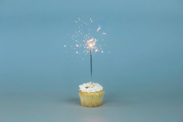 Em frente a um fundo azul claro, um cupcake (bolinho) com creme no topo e uma vela de faíscas acesa.