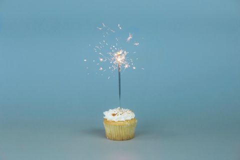 Em frente a um fundo azul claro, um cupcake (bolinho) com creme no topo e uma vela de faíscas acesa.