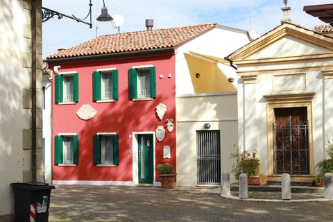 Rua de pedra com duas casas. A da esquerda tem fachada vermelha e possui dois andares, com janelas em verde e branco e telhas marrons no topo; à direita, uma construção amarela com uma grande porta marrom na entrada.