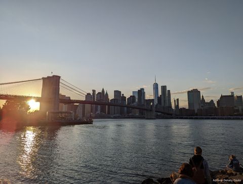Um fim de tarde. Em primeiro plano, um trecho largo de rio com algumas pessoas observando o cenário ao fundo, que conta com uma enorme ponte suspensa e uma linha de prédio, com o sol se pondo atrás.