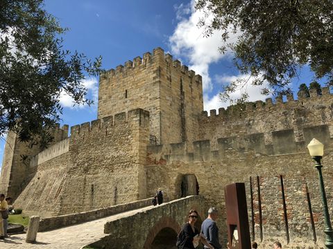 Uma rampa de pedra leva a um castelo de muralhas altas e uma torre quadrada.