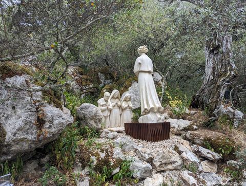 Em uma área rodeada por mata, uma área com pedras possui a estátua de um anjo, com três crianças ajoelhadas rezando logo à sua frente. As esculturas são brancas.