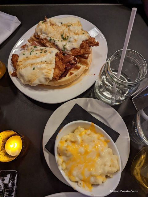 Em uma mesa escura, uma foto de cima mostra dois pratos. Na parte superior, temos waffles coberto com frango, bacon e queijo; na parte inferior, um prato de macarrão com um creme de queijo.