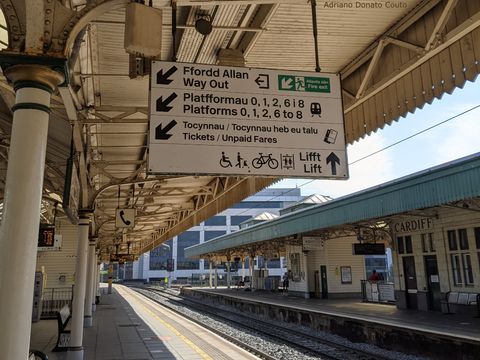 Em uma estação antiga de trem, uma placa branca em primeiro plano exibe informações em inglês e galês.
