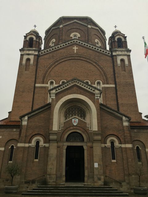 Grande igreja marrom, com detalhes claros em várias áreas. Há uma grande porta preta na frente, com um brazão logo acima, seguido por um arco claro. O céu está cinza, próprio de dia chuvoso.