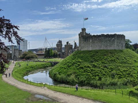 Extensa área gramada, com prédios antigos e novos ao fundo. Muitas pessoas passeiam pela área. À direita, em primeiro plano, um pequeno morro rodeado por um fosso, com as ruínas de um castelo no topo.