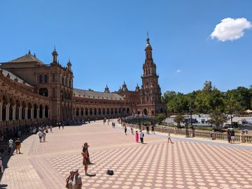 Em primeiro plano, uma via coberta por uma espécie de cerâmica vermelha e alaranjada. Do lado esquerdo, um prédio avermelhado rico em detalhes surge e segue até o fundo da imagem, em um semicírculo. Do lado direito, uma pequena ponte arqueada e uma área verde ao fundo.