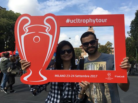 Um rapaz e uma moça morenos seguram uma placa da UEFA Champions League em Milão e sorriem. No meio da placa, uma abertura permite que se veja o rosto dos dois. Ao fundo, várias pessoas caminhando.