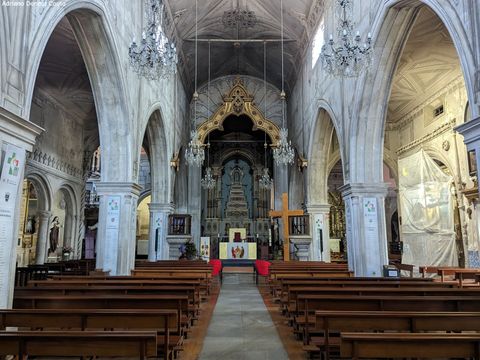 'Vemos o interior de uma igreja a partir da entrada. Os bancos de cada lado são de madeira escura. Ao lado, grandes arcos de pedra e, ao fundo, um altar ricamente ornado.'