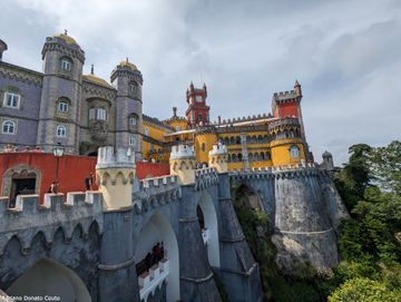 Sob um céu nublado e acinzentado, vemos um palácio com várias torres coloridas e mais de um nível de construção. Abaixo, vemos longas estruturas acinzentadas que sustentam tudo. Há copas de árvore no canto direito inferior.