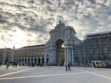 Uma grande praça, com um arco ladeado de prédios antigos logo ao fundo. No topo do arco, uma escultura de uma mulher com os braços abertos e alguns outros elementos. O céu está carregado de nuvens.