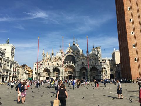 Pessoas e pombos circulam por uma área em frente a uma grande basílica. À direita, vista parcial de um campanário com tons terrosos. Ao centro, três mastros vermelhos com formas douradas na ponta. Logo atrás, a basílica em tons claros e dourados, com cinco grandes portas arqueadas. No topo, há mais um arco para cada porta. Há uma grande cúpula central.
