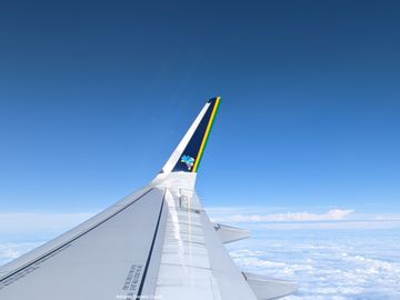 De fundo, temos uma longa área de céu azul, começando de um tom médio no topo até algo próximo de branco, no meio da imagem. Em primeiro plano, parte da asa de um avião, que tem faixas em azul, amarelo e verde em sua ponta, além de um mapa estilizado do Brasil.