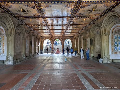Vista de um salão ladeado por arcos ornamentados, com arcos que levam para uma escadaria no fundo. O teto é coberto de azulejos ricamente pintados. Há algumas pessoas no local, incluindo uma senhora com um vestido rosa volumoso.