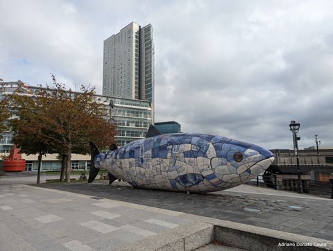 Em uma área de passagem de pedestres à beira de um rio, temos uma pequena árvore e um peixe azul gigante, formado por inúmeros azulejos azuis e brancos.
