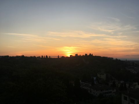 Pôr do sol em tons de laranja e amarelo, refletido em nuvens bem suaves. Na parte inferior da imagem, vê-se levemente alguns prédios, com a silhueta do monte (e de suas árvores) cortando a figura ao meio.
