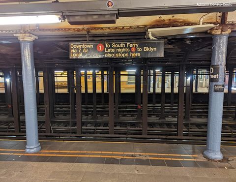 Em primeiro plano, uma plataforma de cimento, com duas colunas acinzentadas. Ao fundo, uma série de colunas de metal separam faixas com trilhos de trem. No topo, uma placa preta com escritos das linhas de metrô daquela plataforma.