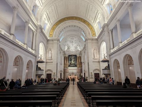 Vemos o interior de uma basílica a partir da entrada. O espaço é ladeado por bancos de madeira escura, com o topo ornamentado com arcos e colunas, tudo em tons claros.