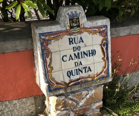 Placa de nome de rua formada por azulejos de fundo branco. Nas bordas da placa, arabescos predominantemente em tons de azul, laranja e roxo. No topo, um brasão. No centro, o nome da rua: Rua do Caminho da Quinta