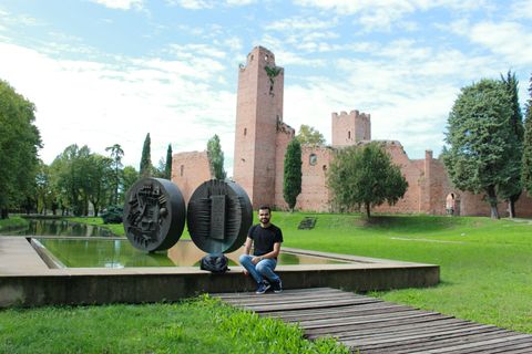 Ao fundo, ruínas de uma espécie de castelo antigo, de cor marrom. Algumas árvores logo em frente. Em primeiro plano, um grande gramado, com um espelho d'água quadrado, com dois discos de metal ornados com relevos. Um rapaz está sentado na borda da frente do espelho d'água.