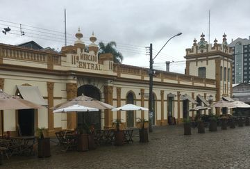 Prédio antigo do Mercado Central de Pelotas. O edifício tem predominantemente tom de amarelo e acabamento em marrom. Em frente, rua de pedra com alguns guarda-sóis. O céu está nublado.