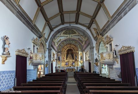 'Vemos uma pequena igreja por dentro, a partir da porta. De paredes brancas, os lados da igreja possuem imagens e outros ornamentos. Ao fundo, um altar dourado cheio de detalhes, com teto pintado.'