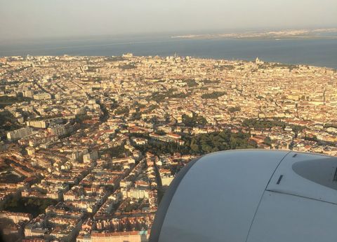 Na parte de baixo, à direita, um pedaço da turbina do avião. Ao fundo, a vista do alto de um mundaréu de construções, todas em tom meio dourado pela luz do sol. Um pouco mais ao fundo, temos o leito largo do rio Tejo.