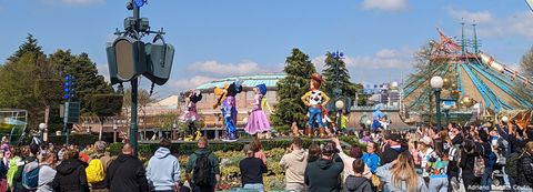 Em uma praça rodeada de pessoas, algumas pessoas fantasiadas de personagens Disney dançam.
