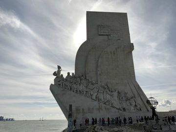Em um monumento em formato de paralelepípedo, uma rampa lateral, que sobe em direção ao rio (à esquerda), possui uma série de esculturas, sendo algumas de navegadores e outras figuras importantes do período das grandes navegações portuguesas. Há também várias pessoas ao redor do monumento e o céu está cheio de nuvens. Este lado fotografado está contra o sol, ou seja, coberto de sombra.