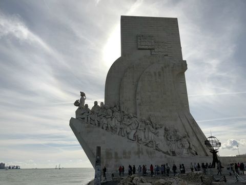Em um monumento em formato de paralelepípedo, uma rampa lateral, que sobe em direção ao rio (à esquerda), possui uma série de esculturas, sendo algumas de navegadores e outras figuras importantes do período das grandes navegações portuguesas. Há também várias pessoas ao redor do monumento e o céu está cheio de nuvens. Este lado fotografado está contra o sol, ou seja, coberto de sombra.