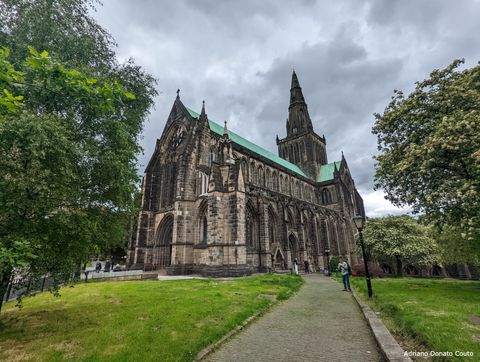 Em uma área gramada e ladeada de árvores, um caminho de pedra leva a uma grande catedral gótica de paredes escurecidas e telhado azul esverdeado. O ângulo de visão é diagonal a partir de sua quina direita.