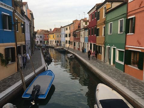Partindo do primeiro plano e indo em direção ao fundo, um canal com alguns barcos parados em suas margens.O canal é margeado por uma série de pequenos prédios de dois andares, cada um com fachada de uma cor diferente. Marca registrada de Burano (Veneza).