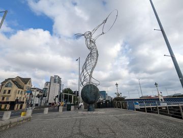 Em uma calçada de pedras, uma escultura feita de tubos metálicos representa uma figura feminina, que segura um anel de metal.
