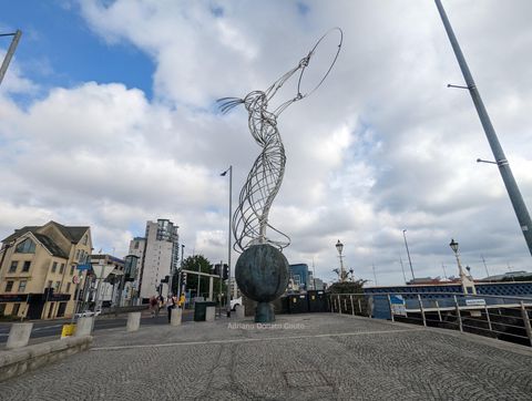 Em uma calçada de pedras, uma escultura feita de tubos metálicos representa uma figura feminina, que segura um anel de metal.