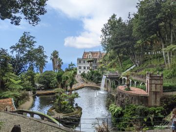 Em primeiro plano, uma lagoa com cascata artificial, cercada por plantas de diversos tipos; em segundo plano, um imponente casarão de 3 andares, com telhado todo decorado. Ao fundo, o céu azul se mistura com o mar.